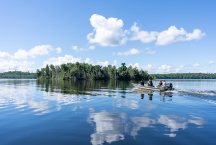 A motorboat cruises by a small island on a bright, sunny day with the cloud reflecting in the rippling water.