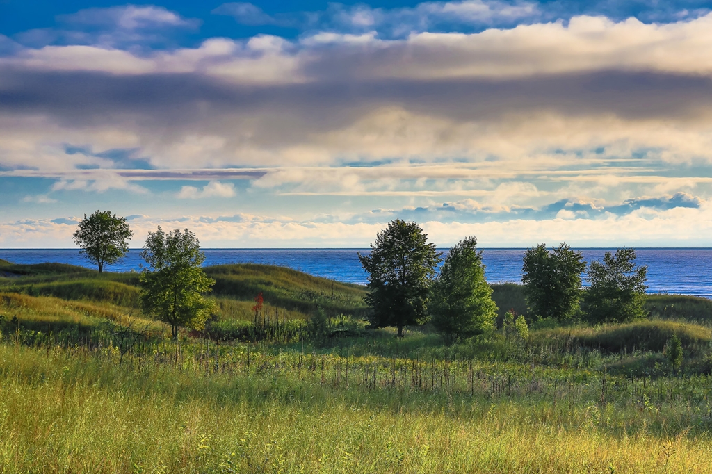 Photograph of rolling green dunes on a shore overlooking blue water and under a cloud-filled sky. Photo is "Early Morning Dew” by Matthew John George and was submitted for the 2015 Great Waters Photo Contest.