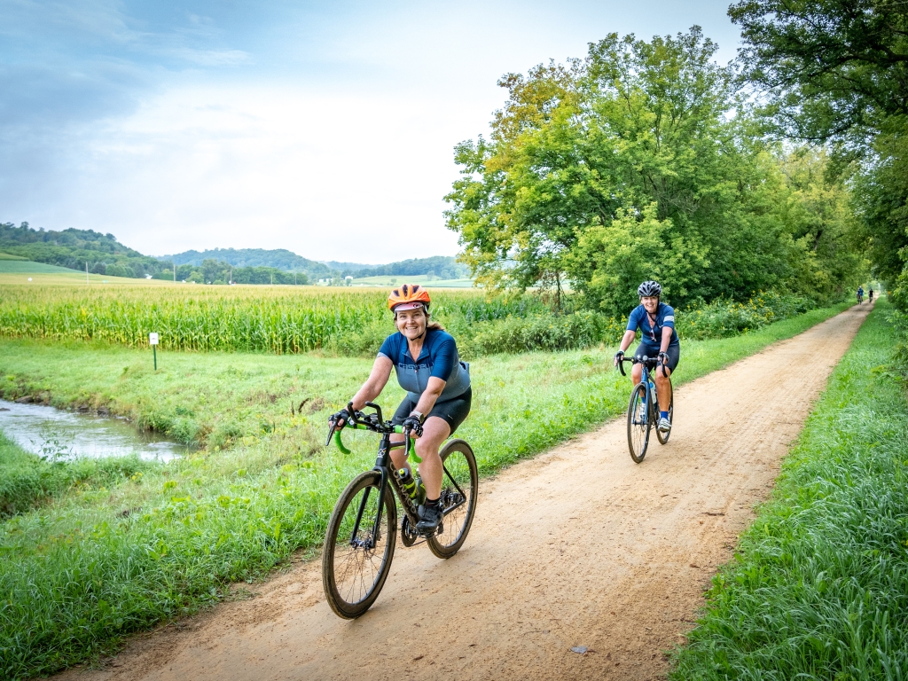 Two happy bikers travel on a gravel road along fields and trees.